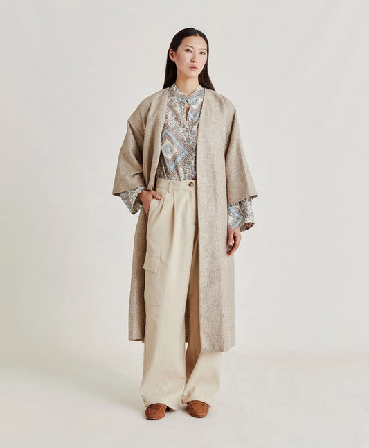 A woman poses against a plain light background wearing the MOMONI - CALL Lightweight Coat in Lurex Basketweave over a patterned shirt and loose, high-waisted beige wide-leg pants. She has straight dark hair and brown shoes.