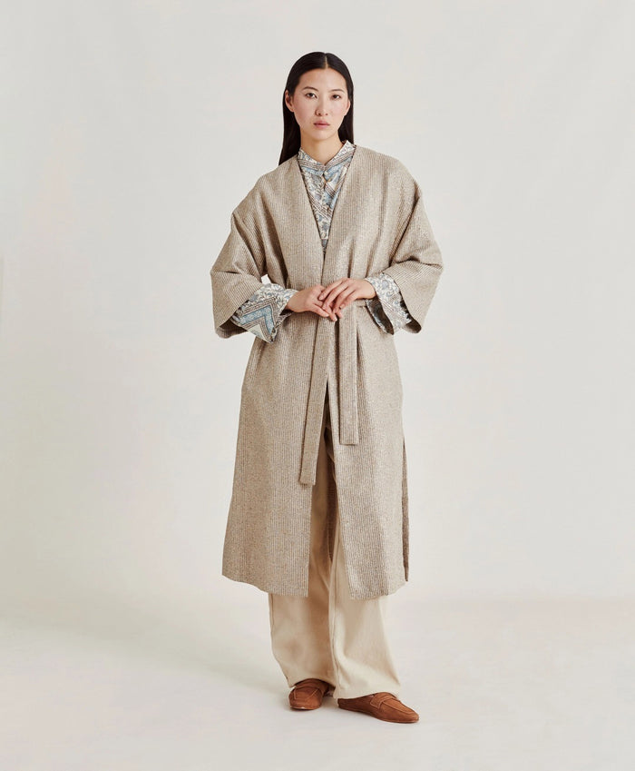 A woman poses against a plain light background wearing the MOMONI - CALL Lightweight Coat in Lurex Basketweave over a patterned shirt and loose, high-waisted beige wide-leg pants. She has straight dark hair and brown shoes.