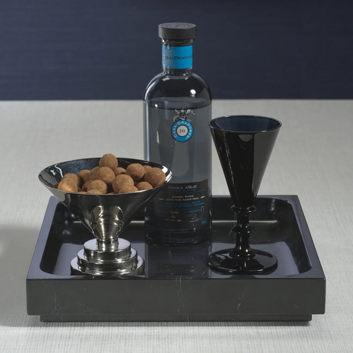 A black bottle of tequila sits on a ZODAX NERO MARQUINA NATURAL MARBLE TRAY - LARGE, accompanied by a black goblet and bowl of chocolate treats, all arranged on a light surface with a dark backdrop.