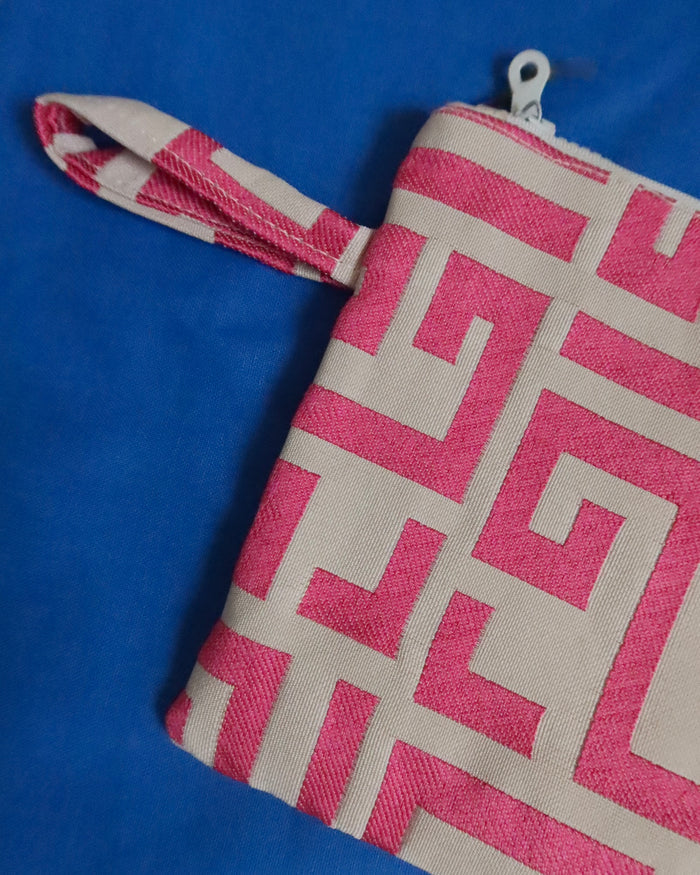 A small zip pouch with a wrist strap, featuring a pink and off-white geometric pattern, shown on a solid blue background.