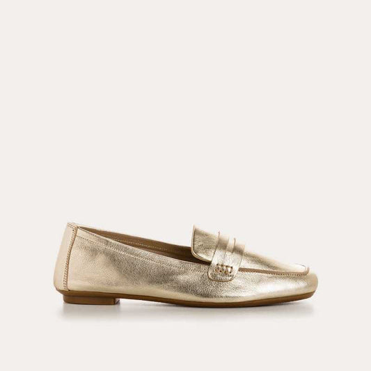 The REQINS - HEMA Metallic Loafer in Platinum features shimmering leather, a low heel, and a decorative front strap, showcased on a plain white background.