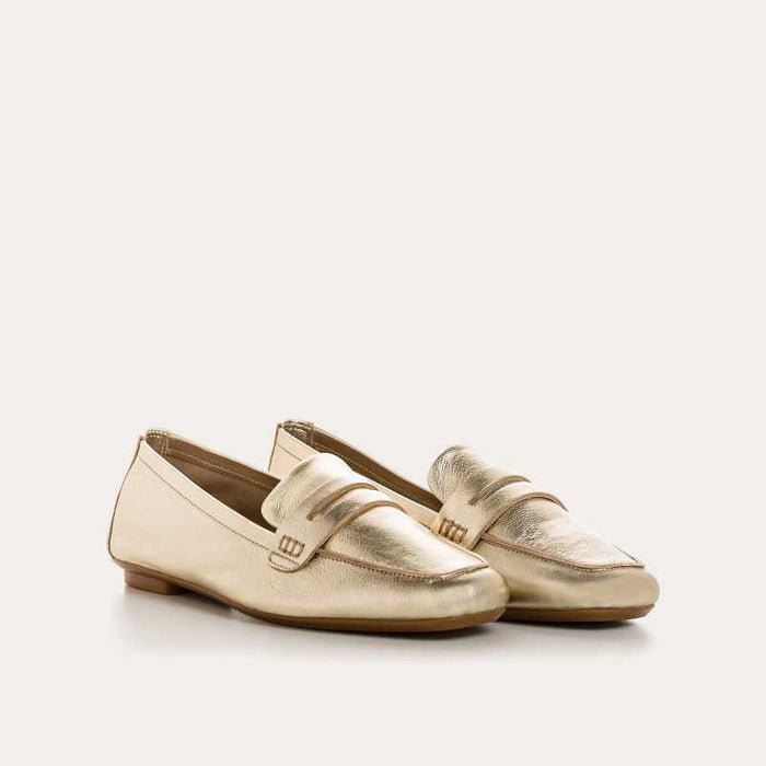 The REQINS - HEMA Metallic Loafer in Platinum features shimmering leather, a low heel, and a decorative front strap, showcased on a plain white background.