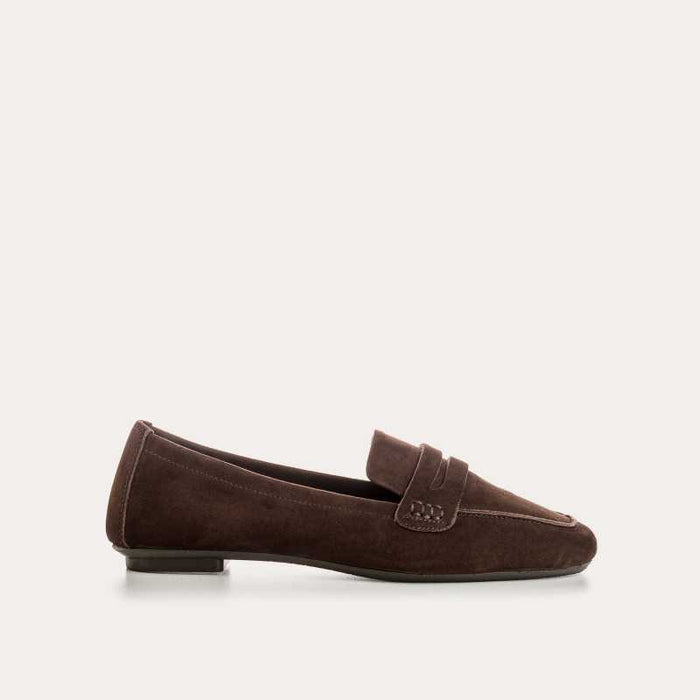 A person stands on a light floor wearing rolled-up blue jeans and REQINS HEMA suede loafers in Marron, showcasing the artisanal craftsmanship of REQINS.