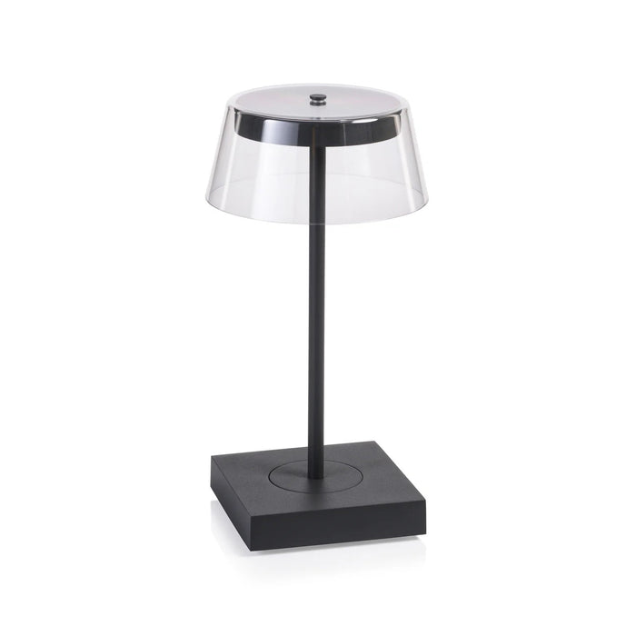 The SARDINIA RECHARGEABLE TABLE LIGHT by ZODAX, with a transparent shade and sleek dark base featuring adjustable brightness, is illuminated on a textured surface beside the book "SHIRO TSUJIMURA: An Art of Living.