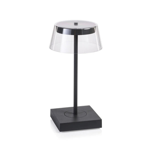 The SARDINIA RECHARGEABLE TABLE LIGHT by ZODAX, with a transparent shade and sleek dark base featuring adjustable brightness, is illuminated on a textured surface beside the book "SHIRO TSUJIMURA: An Art of Living.
