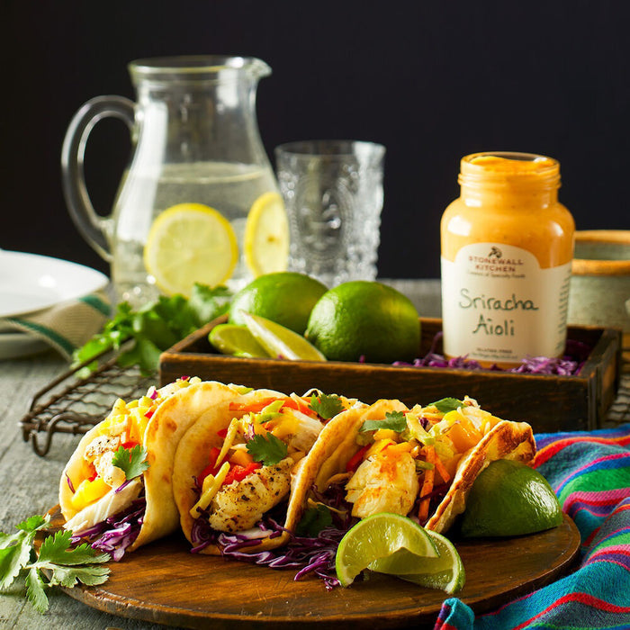 A jar of STONEWALL KITCHEN-SRIRACHA AIOLI features a green lid and beige label with product and brand name, marked "Gluten Free." Inside is creamy, orange, spicy mayo.