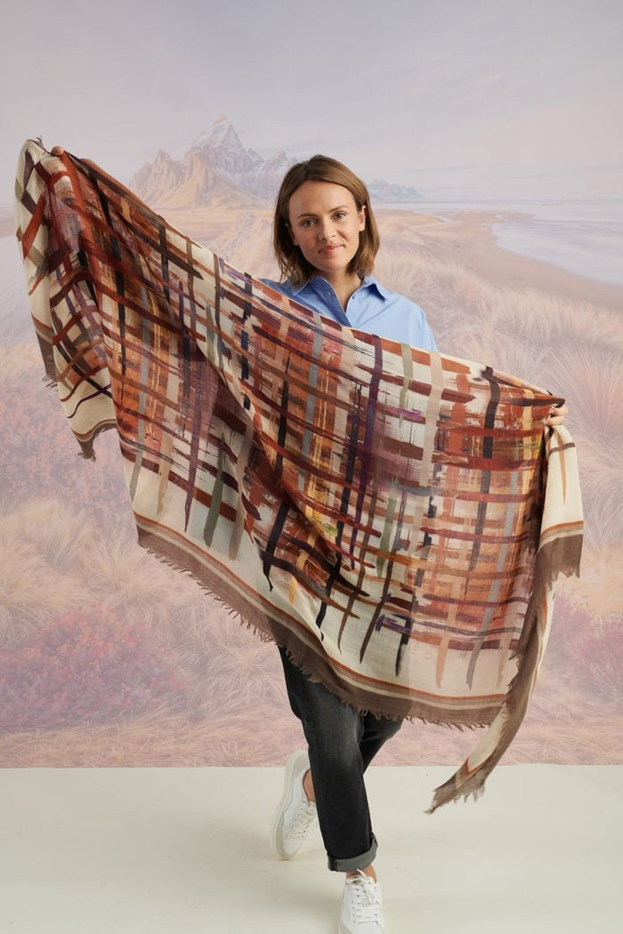 A woman in a blue shirt and jeans stands holding the STORIATIPIC - JULIE WOOL SCARF, a luxury plaid accessory in earthy tones. Behind her, mountains, beach, and grassy dunes create a scenic backdrop.