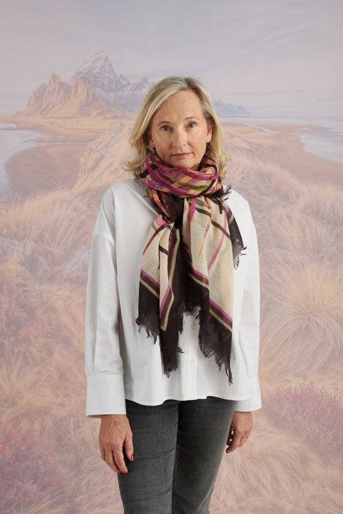 A woman in a blue shirt and jeans stands holding the STORIATIPIC - JULIE WOOL SCARF, a luxury plaid accessory in earthy tones. Behind her, mountains, beach, and grassy dunes create a scenic backdrop.