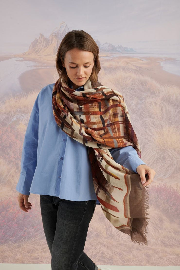 A woman in a blue shirt and jeans stands holding the STORIATIPIC - JULIE WOOL SCARF, a luxury plaid accessory in earthy tones. Behind her, mountains, beach, and grassy dunes create a scenic backdrop.
