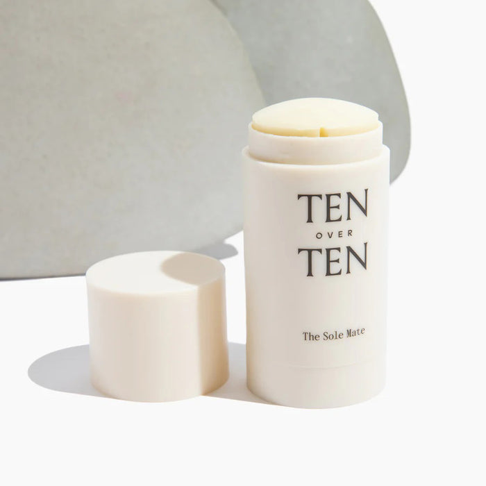 A white tube of TEN OVER TEN - THE SOLE MATE antifungal foot balm sits with its cap off, showing the moisturizer inside, on a light surface against a soft, abstract background.