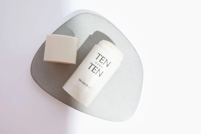 A white tube of TEN OVER TEN - THE SOLE MATE antifungal foot balm sits with its cap off, showing the moisturizer inside, on a light surface against a soft, abstract background.