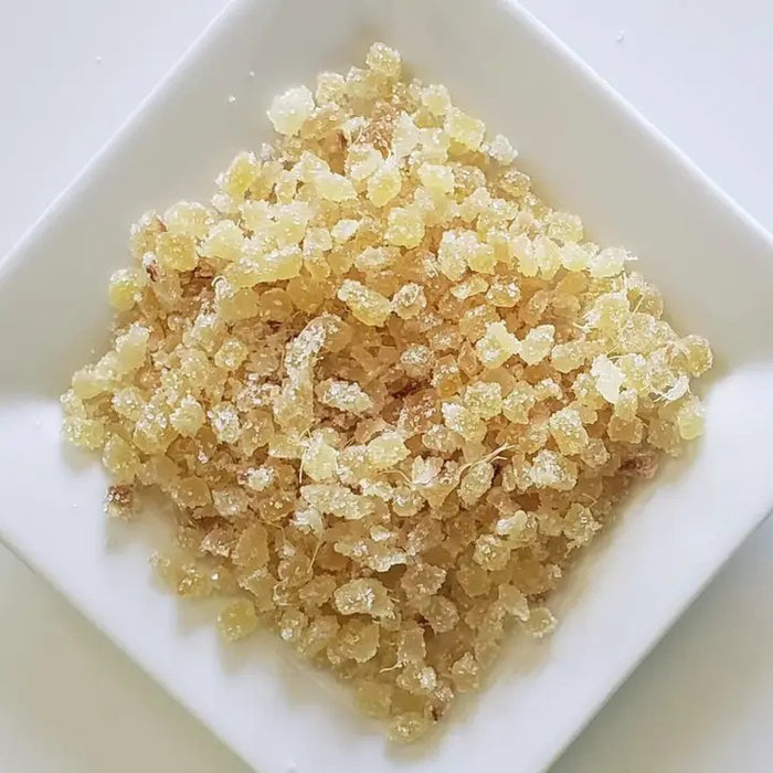 A close-up image of a square white plate containing small, beige-colored, crystal-like granules that appear to be THE CHAI BOX - CANDIED GINGER. The granules have a slightly translucent appearance.