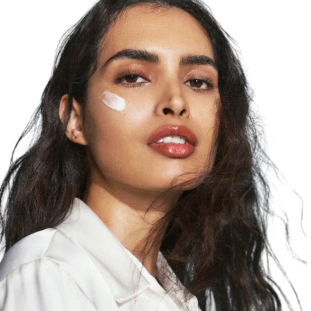 A woman with long dark hair and dewy skin wears a white top and has a streak of U BEAUTY THE SUPER HYDRATOR 50ML on her cheek. She gazes confidently at the camera against a plain white background, highlighting intense hydration.