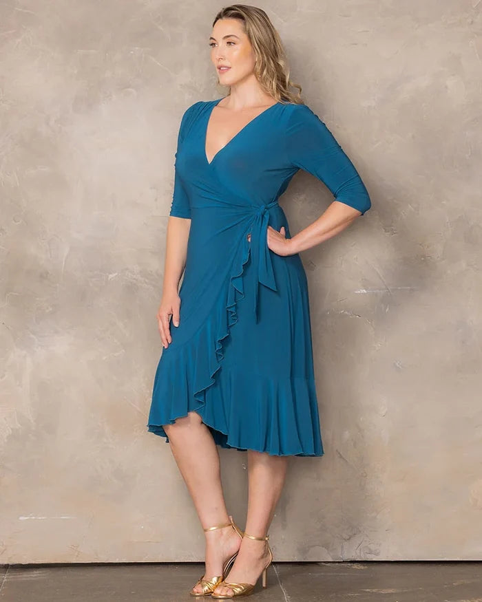 Whimsy Wrap Dress - Sale! in Aegean Blue. Model in a blue ruffle wrap dress and gold heels in front of a beige background.