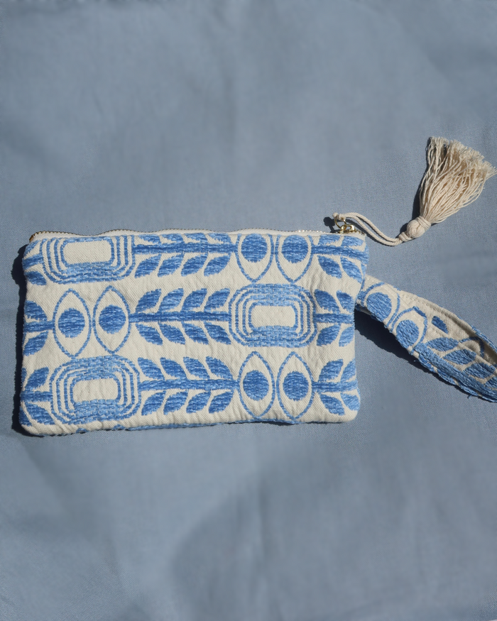 A small woven clutch or pouch with a blue and off-white geometric pattern with evil eye print and olive branches, featuring a wrist strap and a cream tassel zipper pull, laid flat on a blue fabric background. Marabu Greek brand at Aegean Essence