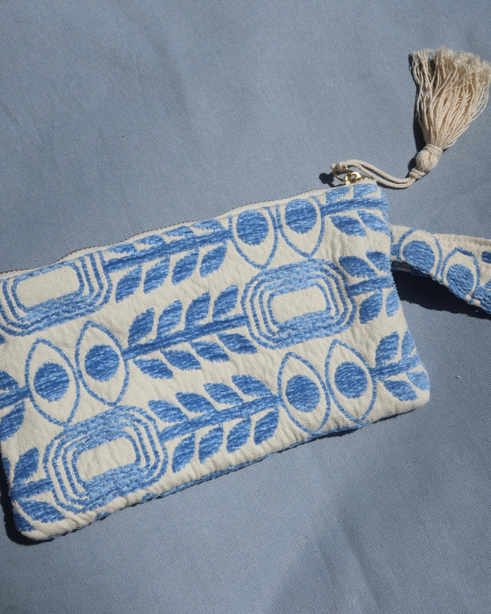 A small woven clutch or pouch with a blue and off-white geometric pattern with evil eye print and olive branches, featuring a wrist strap and a cream tassel zipper pull, laid flat on a blue fabric background. Marabu Greek brand at Aegean Essence