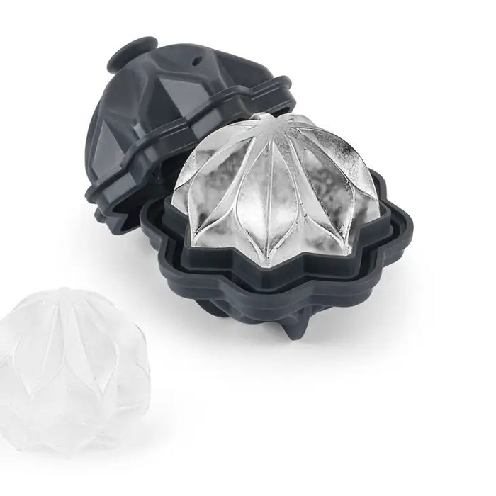 A black W&P - RIPPLE COCKTAIL ICE MOLD by W&P DESIGN, partially open, displays a decorative wave-pattern ice sphere that melts slowly. Another faceted sphere sits nearby on the white surface.