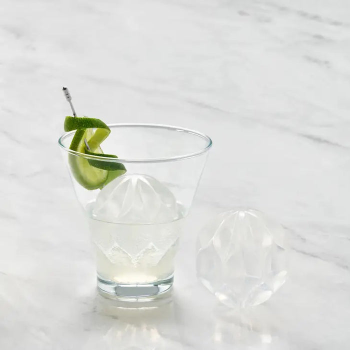 A black W&P - RIPPLE COCKTAIL ICE MOLD by W&P DESIGN, partially open, displays a decorative wave-pattern ice sphere that melts slowly. Another faceted sphere sits nearby on the white surface.
